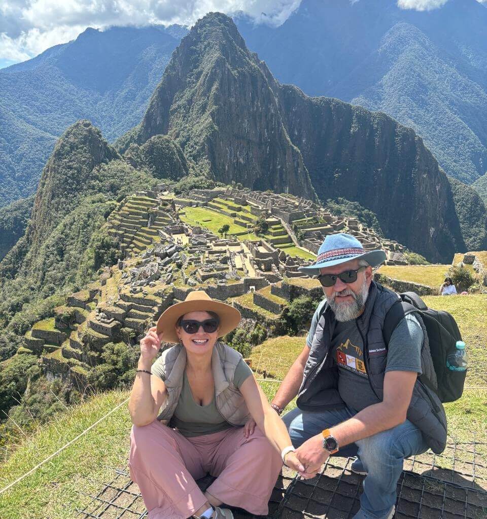 Couple sitting at viewpoint overlooking Machu Picchu terraces and Huayna Picchu mountain under partly cloudy August sky.