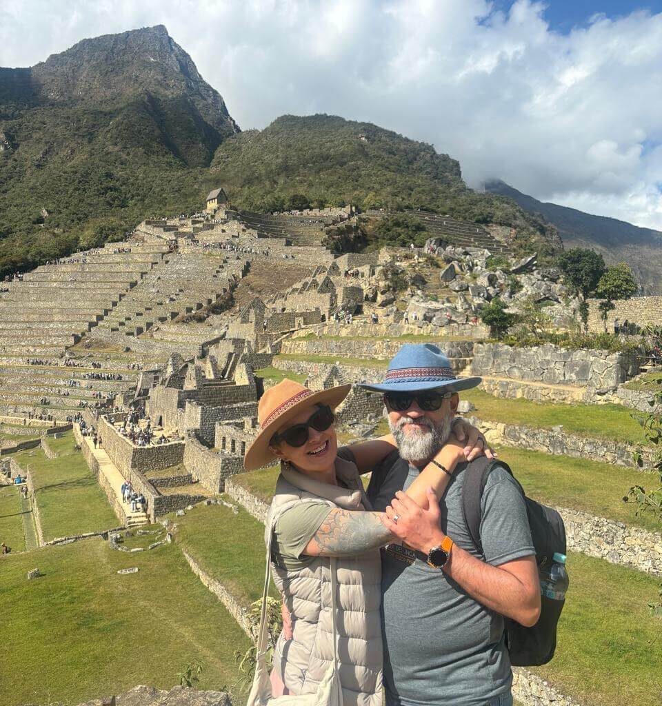 Couple hugging with Machu Picchu ruins and surrounding Andes mountains visible in August daylight.