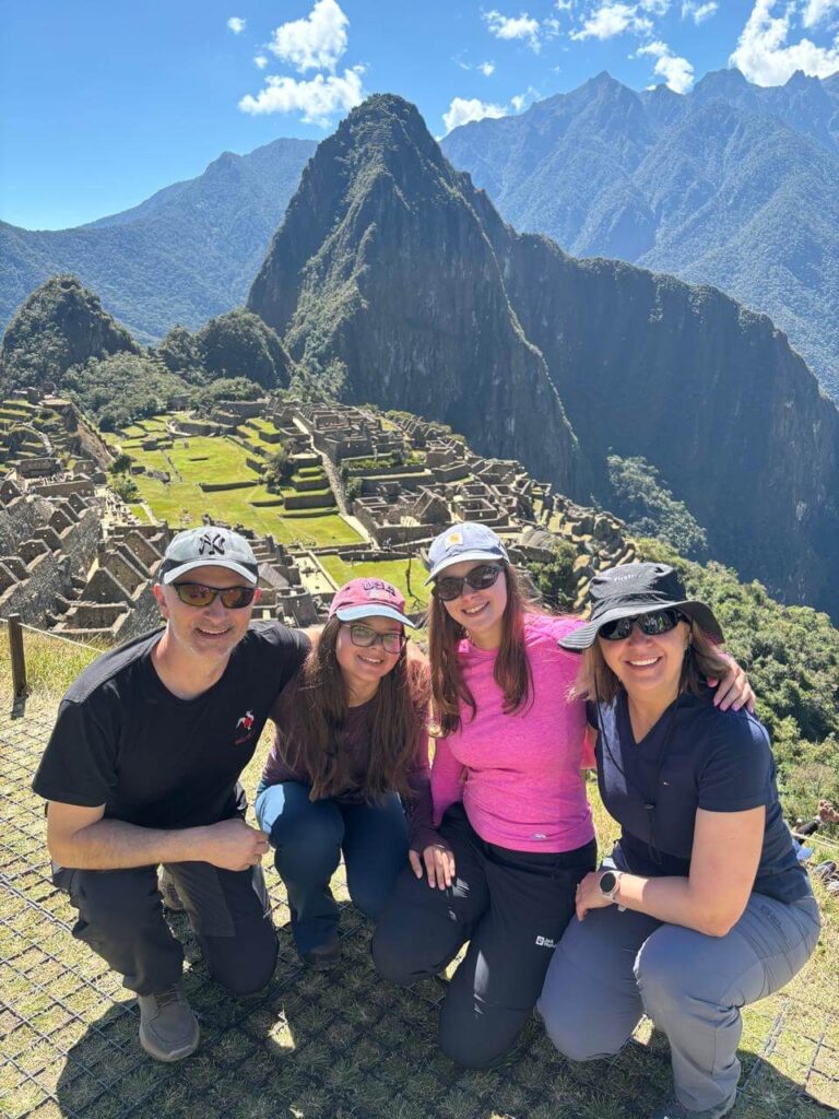 Family group posing at Machu Picchu viewpoint with clear mountain visibility and bright August sunlight.