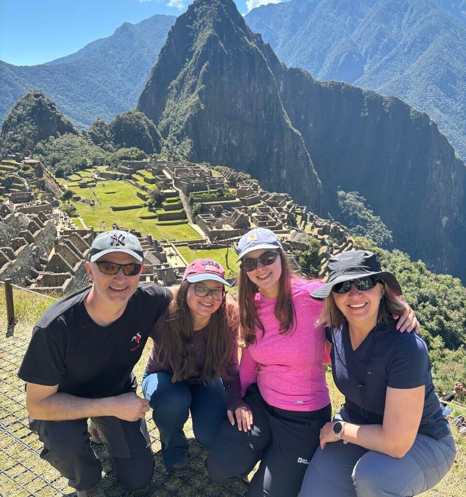 Family group posing at Machu Picchu viewpoint with clear mountain visibility and bright August sunlight.