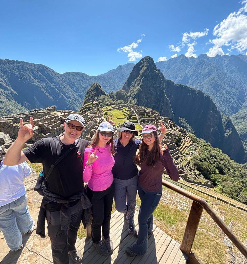 Group of travelers posing at Machu Picchu viewpoint with blue sky and Andes mountains in August.