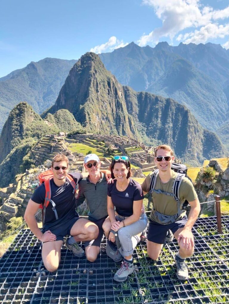 Group of hikers posing at Machu Picchu viewpoint with ruins and Andes mountains under blue August sky.