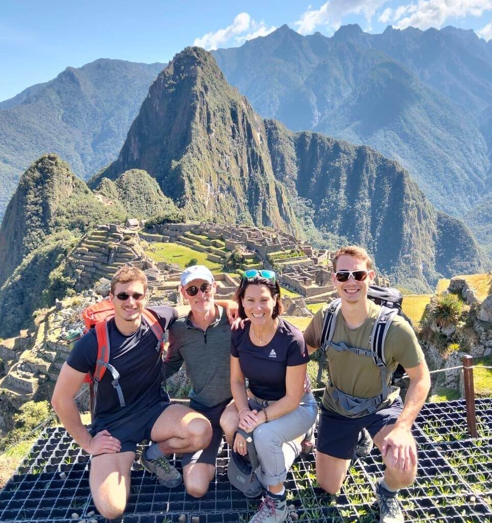 Group of hikers posing at Machu Picchu viewpoint with ruins and Andes mountains under blue August sky.
