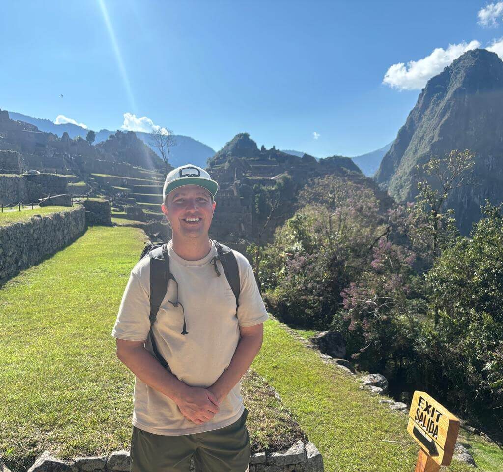 Solo traveler standing near stone terraces at Machu Picchu with mountains and blue sky in August.