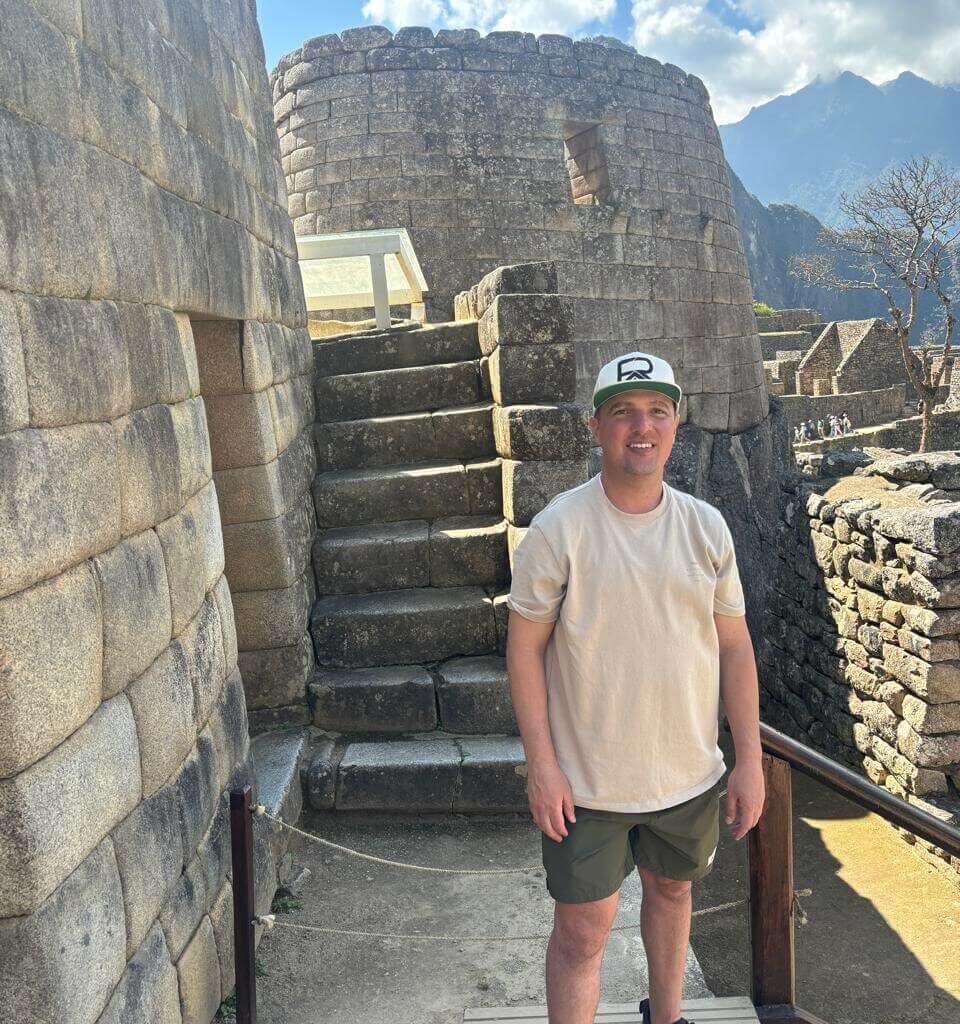Traveler standing next to circular Inca stone structure and stairs inside Machu Picchu on a sunny August day.