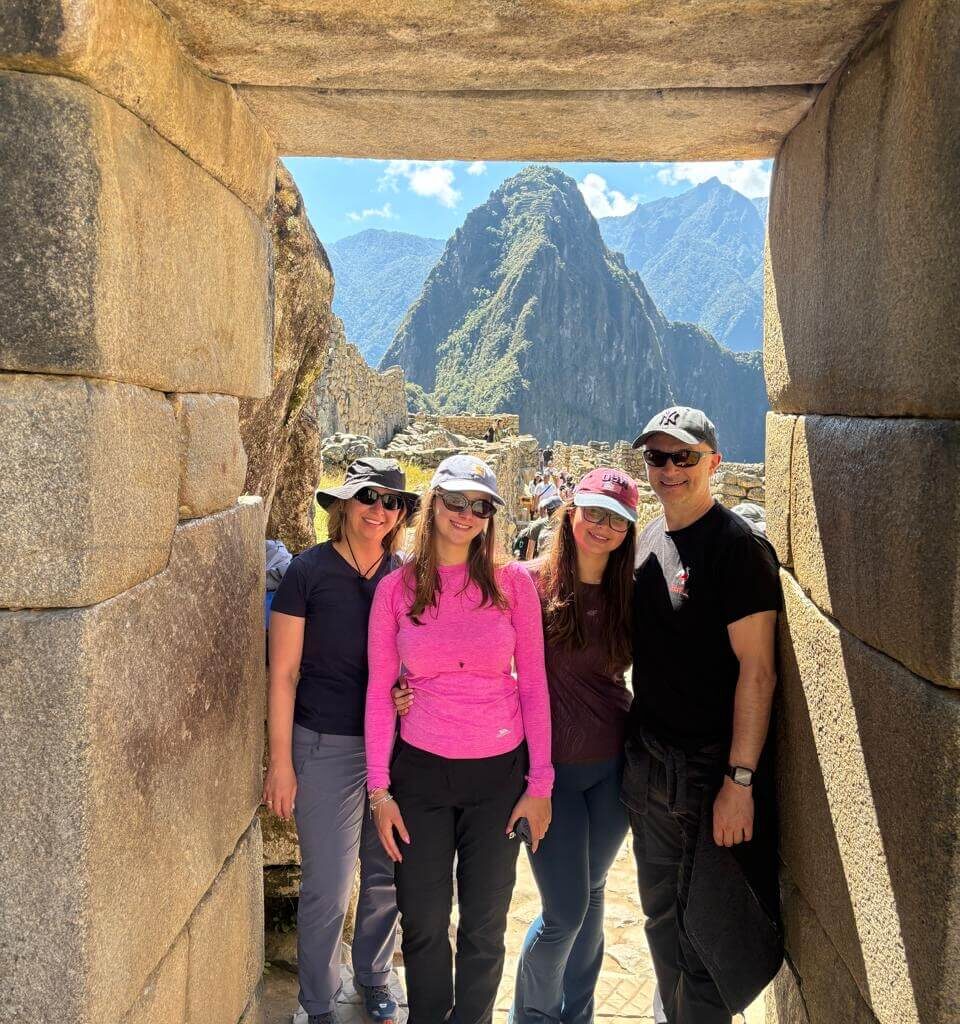 Group of travelers standing inside Inca stone doorway with Machu Picchu ruins and Huayna Picchu mountain in August.