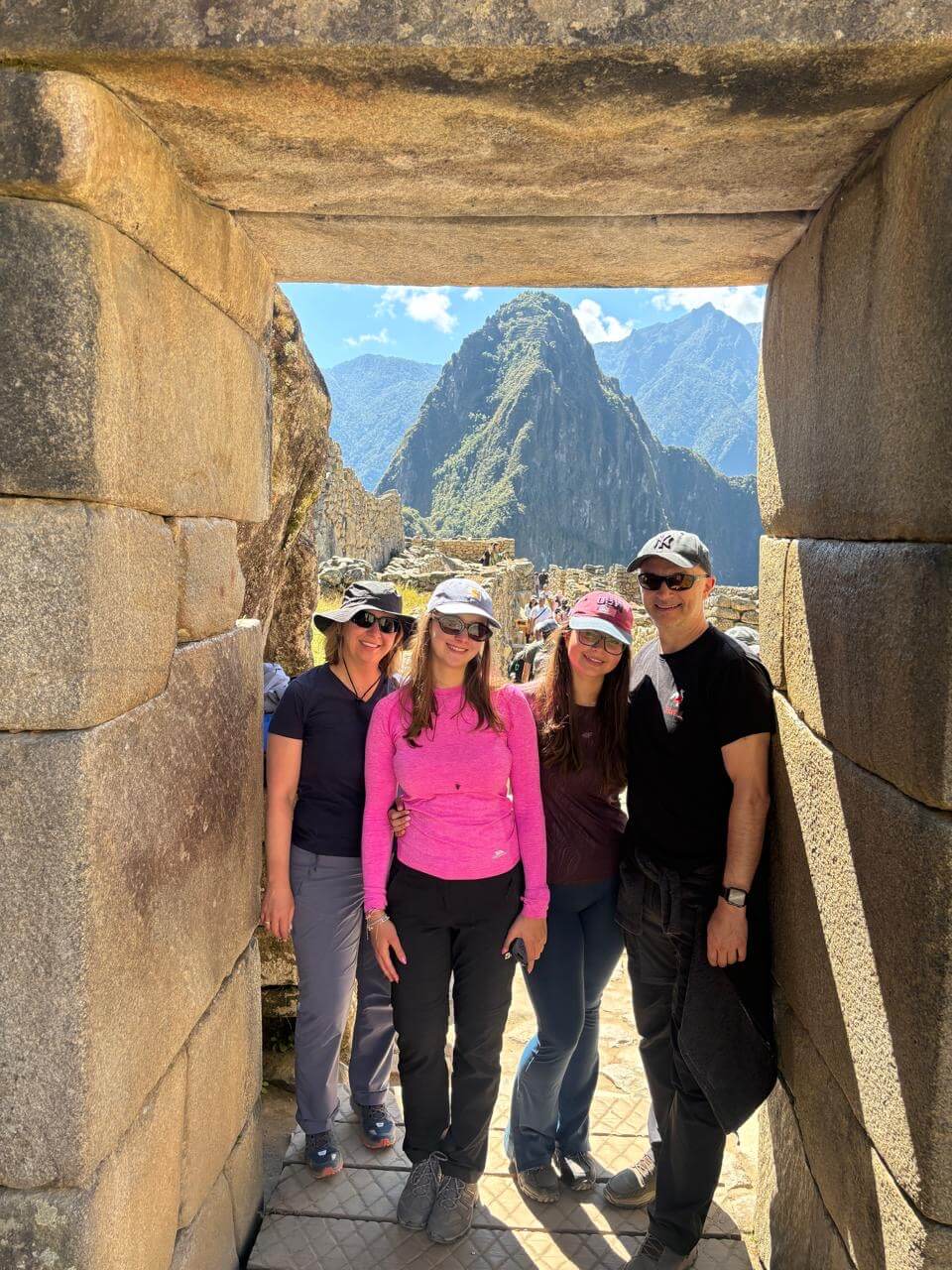 Group of travelers standing inside Inca stone doorway with Machu Picchu ruins and Huayna Picchu mountain in August.