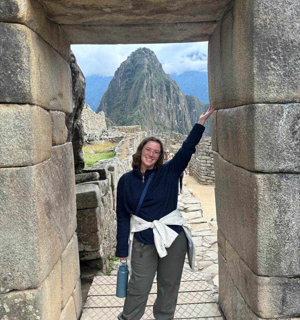 Traveler posing inside Inca stone doorway framing Huayna Picchu mountain and Machu Picchu ruins in August.