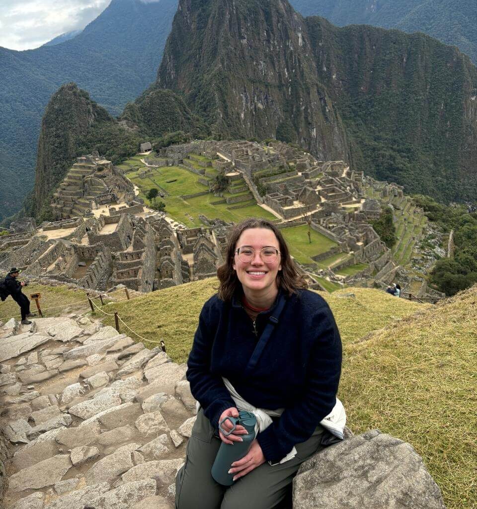 Traveler sitting on stone steps overlooking Machu Picchu ruins and terraces under cloudy August sky.