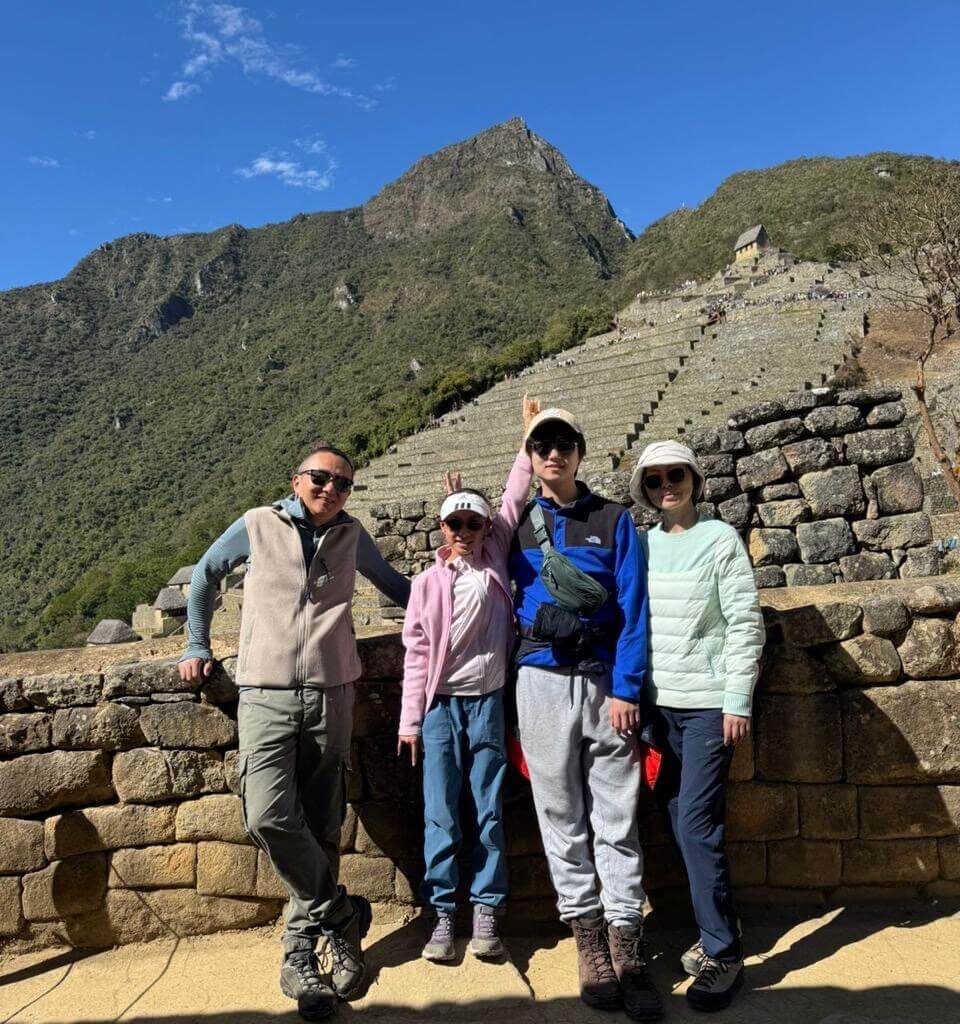 Family standing near stone wall and agricultural terraces at Machu Picchu with mountain backdrop in August.