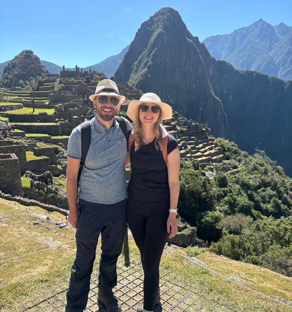 Couple standing at Machu Picchu viewpoint wearing hiking clothes and hats with Huayna Picchu mountain and ruins in background.