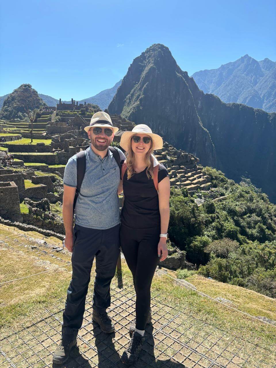 Couple standing at Machu Picchu viewpoint wearing hiking clothes and hats with Huayna Picchu mountain and ruins in background.