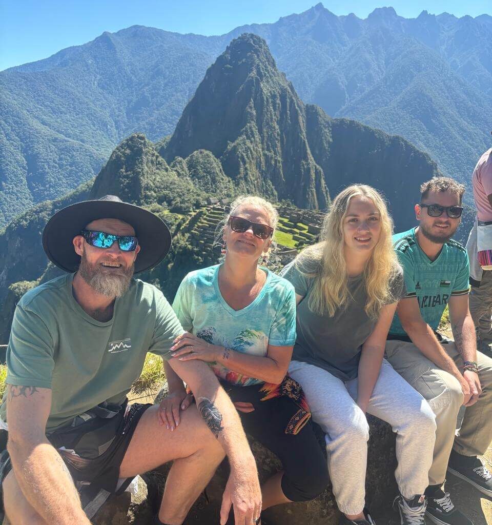 Four travelers sitting on a stone ledge at Machu Picchu with green terraces and Andean mountains visible in July.
