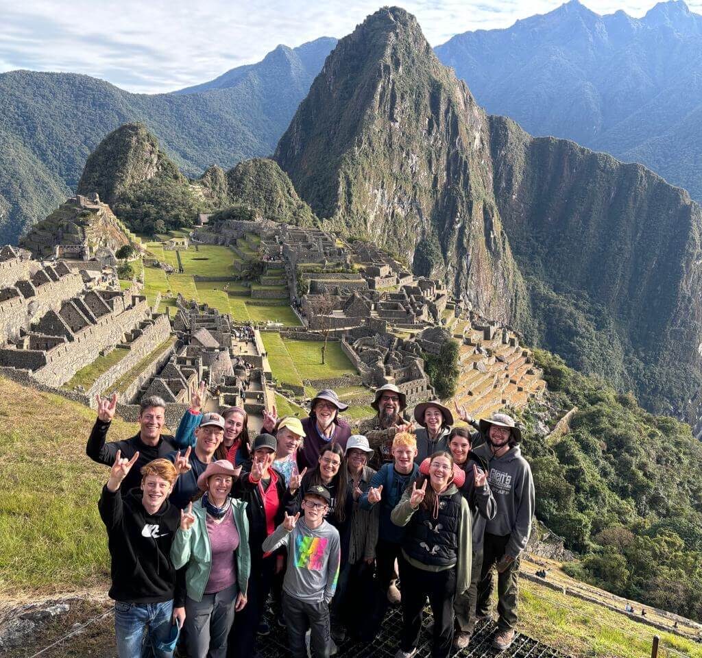 Group of travelers posing at Machu Picchu viewpoint with stone terraces and Huayna Picchu mountain behind under clear July sky.