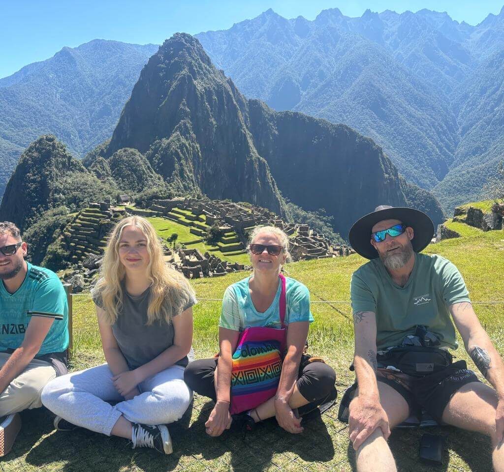 Four people sitting on grass overlooking Machu Picchu terraces and Huayna Picchu mountain under blue July sky.