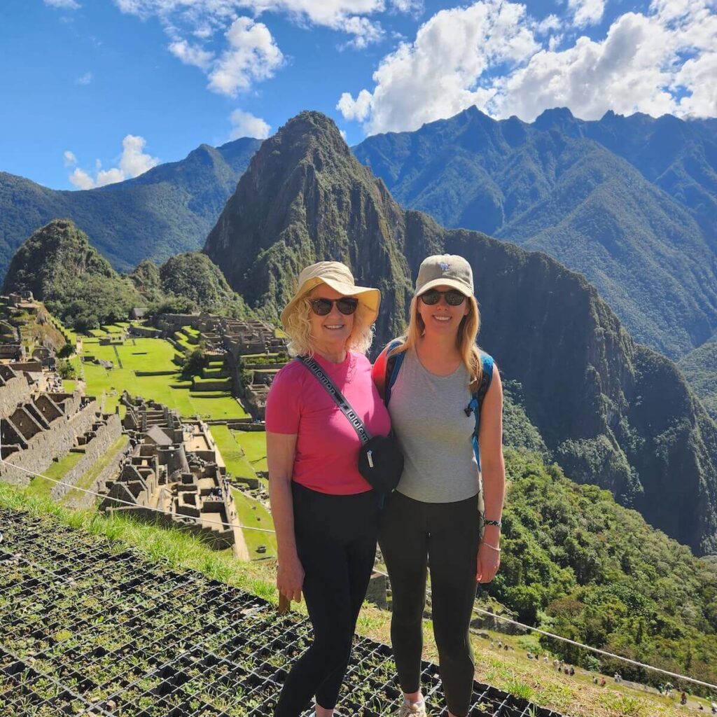 Two travelers posing at Machu Picchu viewpoint with ruins and Huayna Picchu mountain visible on a bright July day.