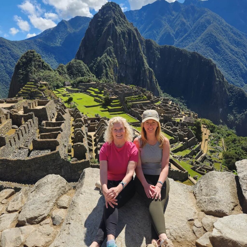 Two travelers sitting on rocks overlooking Machu Picchu ruins and terraces with Andean mountains and clouds in July.