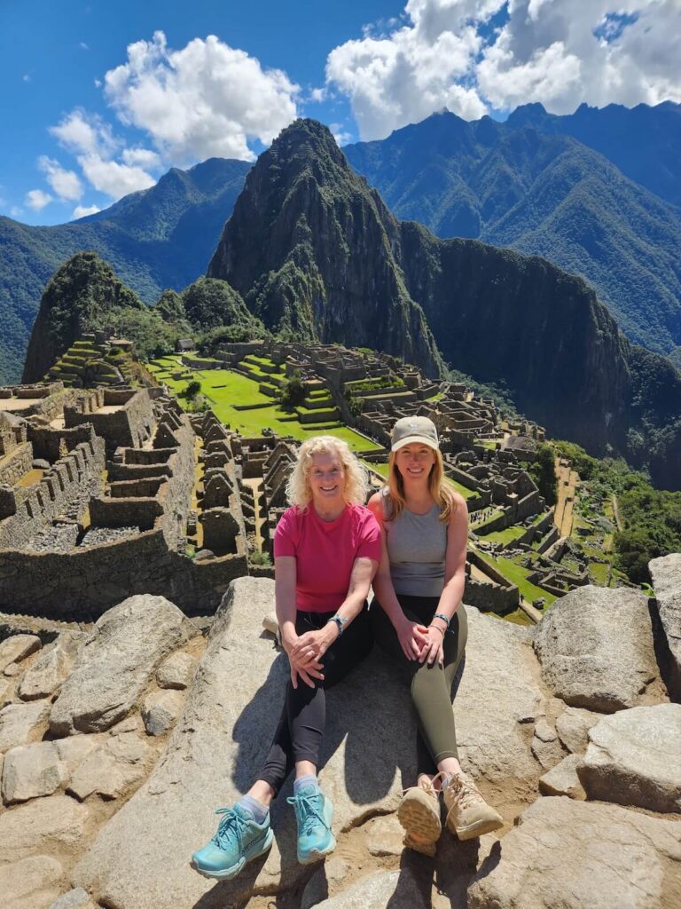 Two travelers sitting on rocks overlooking Machu Picchu ruins and terraces with Andean mountains and clouds in July.