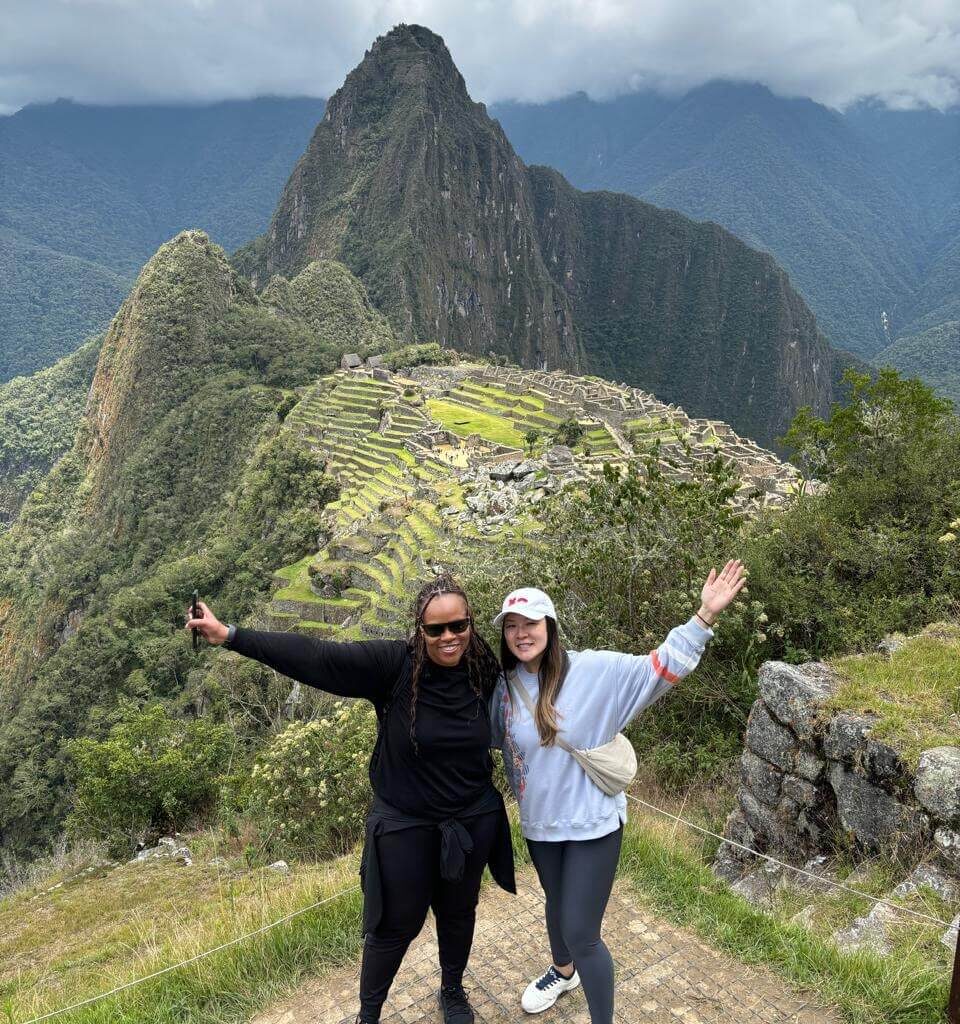 Two travelers posing with Machu Picchu terraces and mountains during October cloudy season.