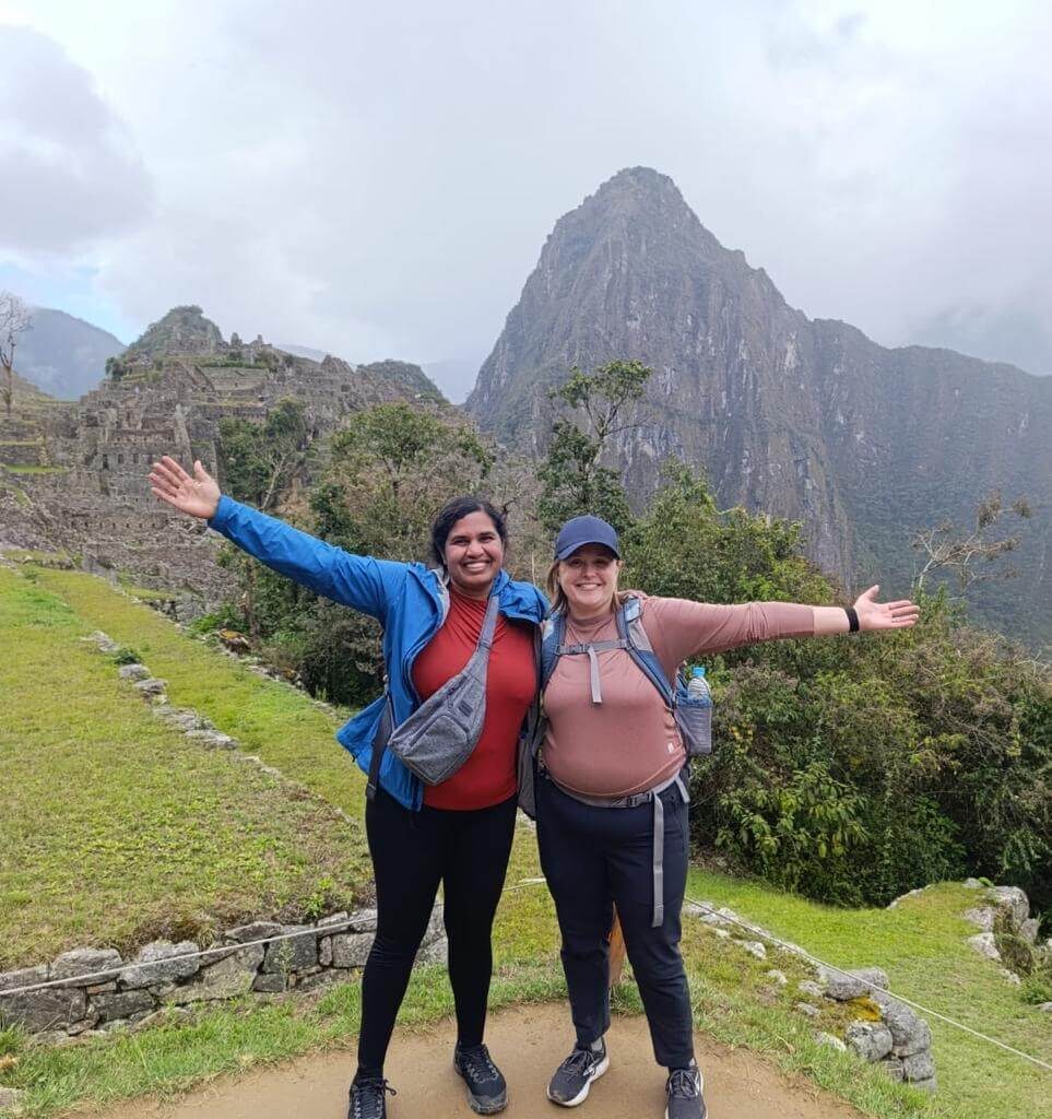 Two hikers posing with arms out at Machu Picchu viewpoint with mountains and clouds in October.