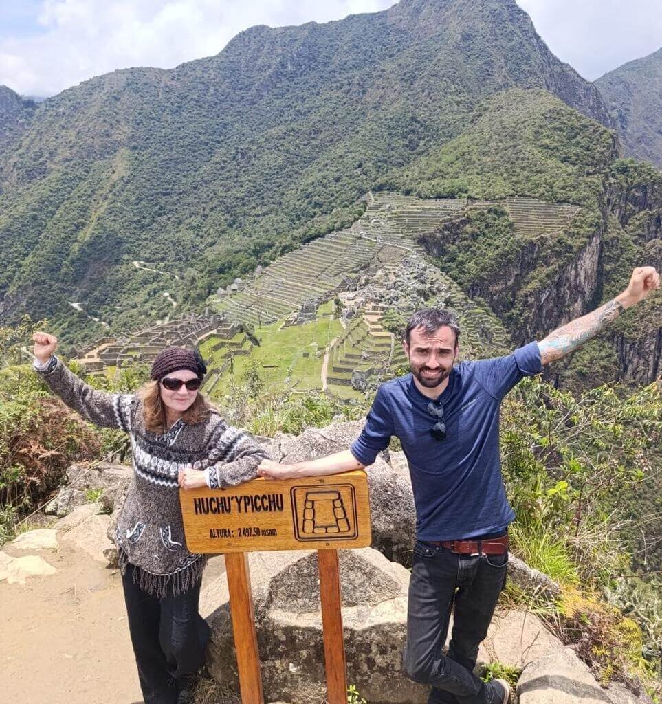 Travelers standing next to Huchuy Picchu sign with Machu Picchu terraces and mountains behind.