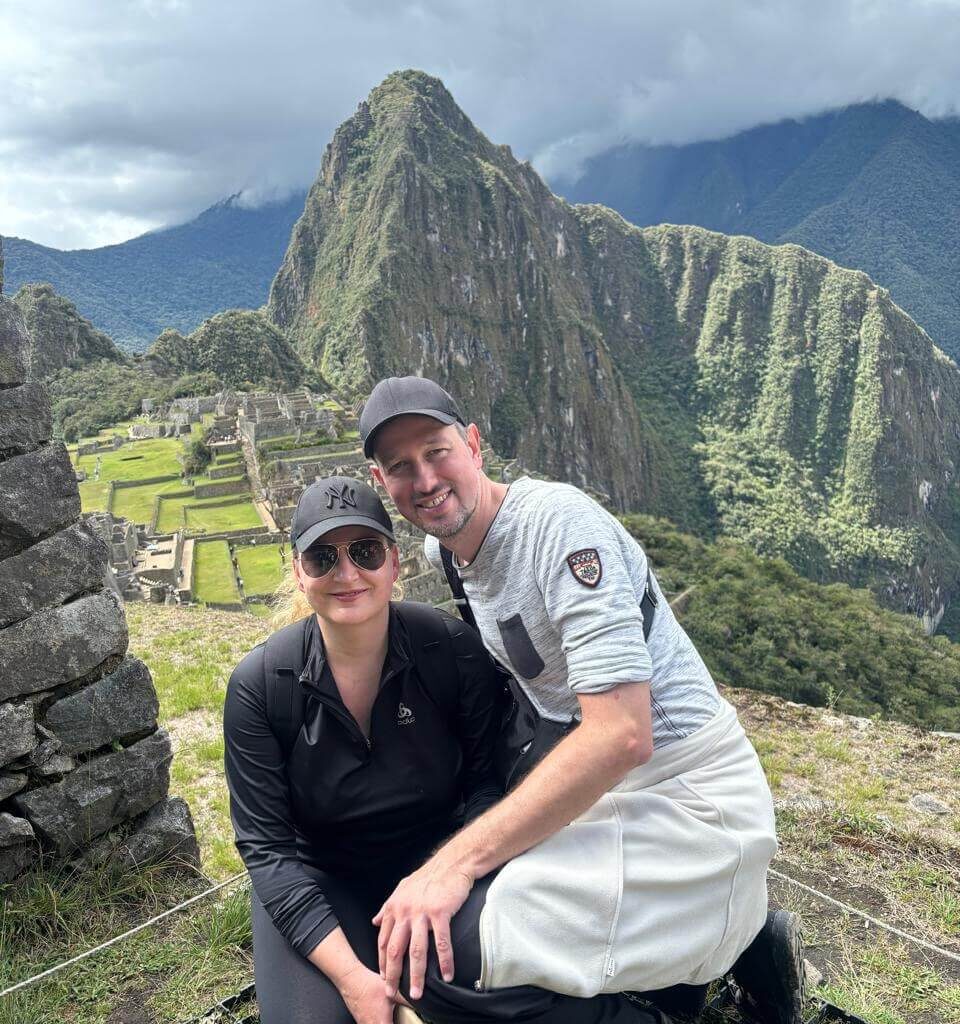 Couple posing at Machu Picchu viewpoint with Huayna Picchu and green terraces behind.