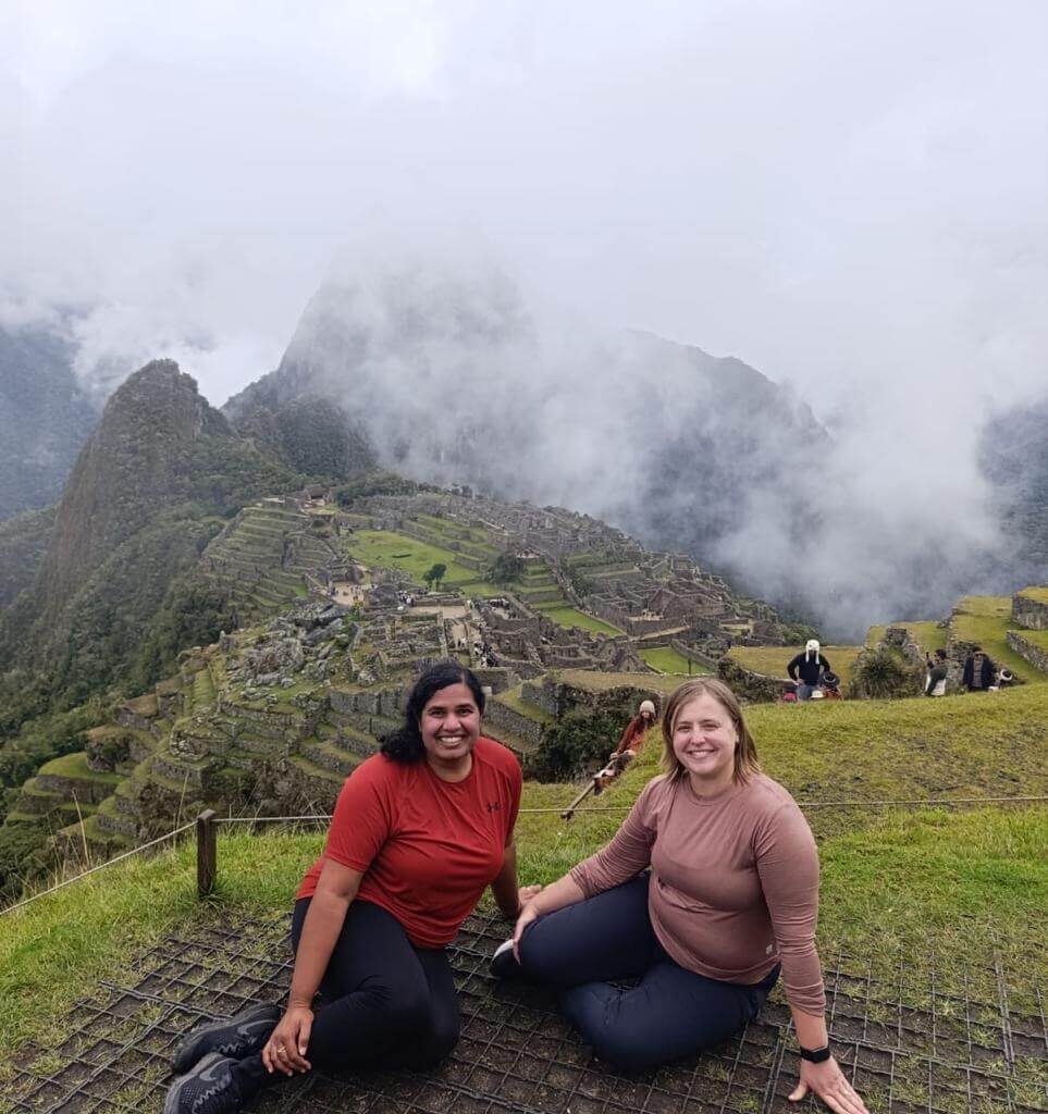Two travelers sitting at Machu Picchu viewpoint with fog covering mountains in October.