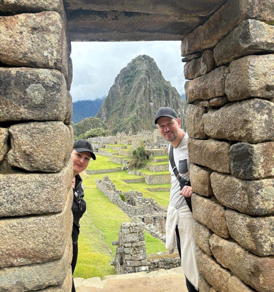 Two travelers peeking through Machu Picchu stone doorway with ruins and green terraces.