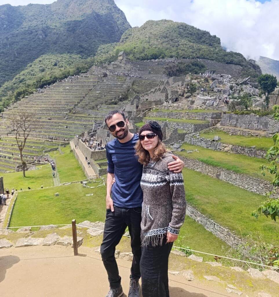 Couple standing above Machu Picchu terraces with blue sky and mountain background.