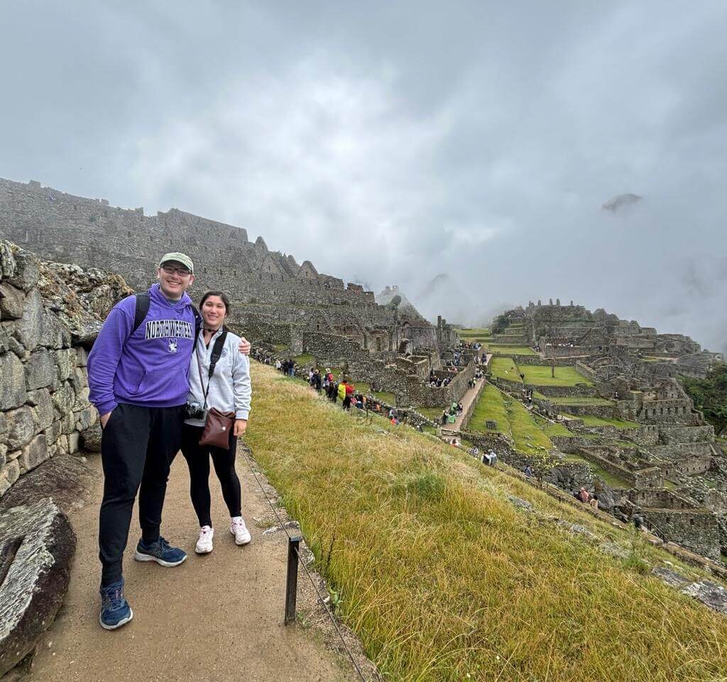 Two travelers standing along Machu Picchu terrace pathway with fog rolling over ruins.