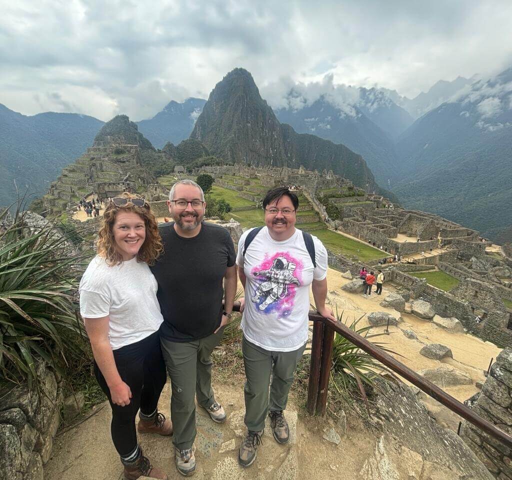 Three visitors posing above Machu Picchu ruins with Andes mountains and cloudy sky in September.