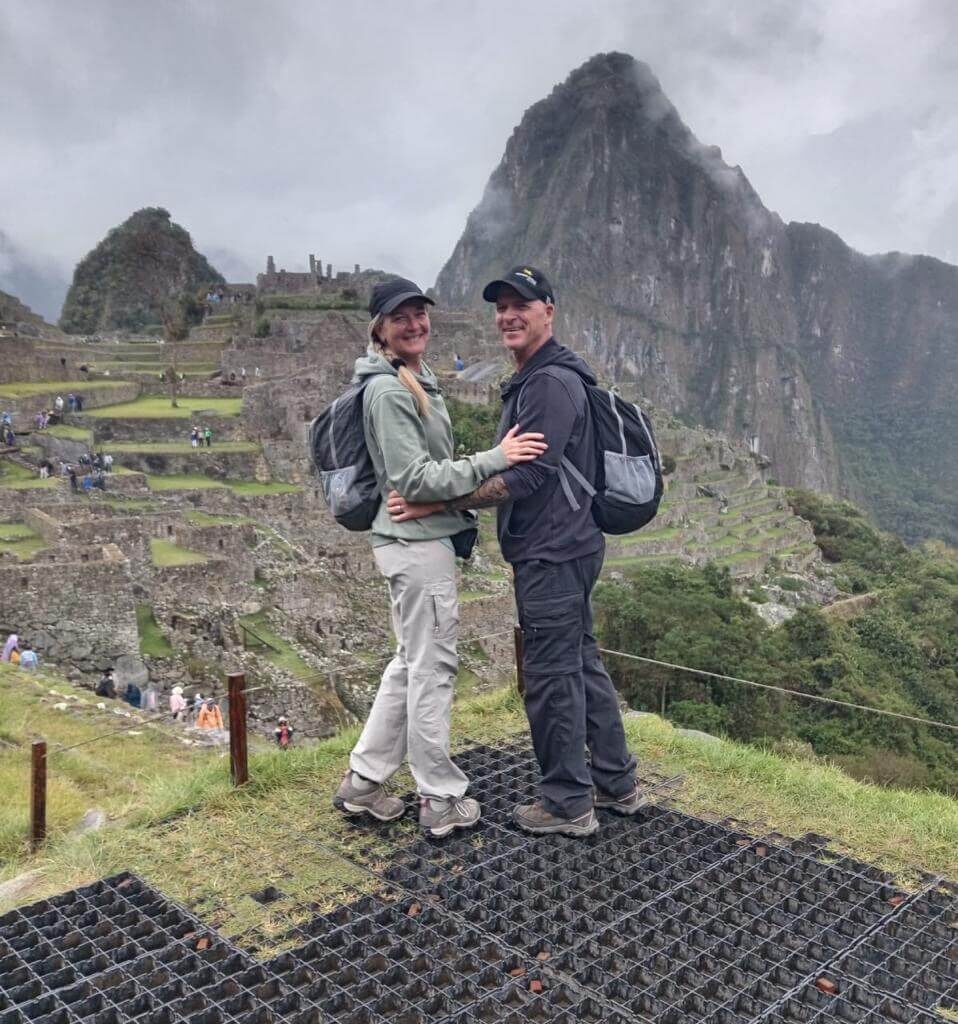 Couple standing at Machu Picchu viewpoint with Inca terraces and Huayna Picchu mountain in background.