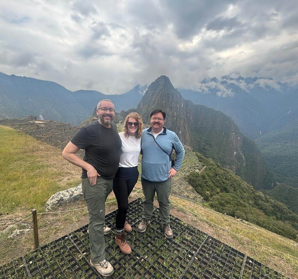 Three travelers standing at a Machu Picchu viewpoint with Huayna Picchu mountain and cloudy Andes landscape in September.