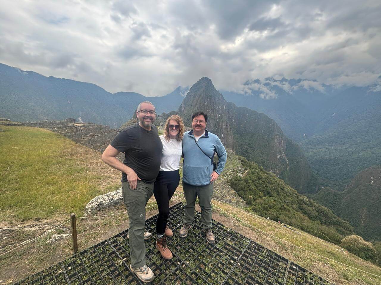Three travelers standing at a Machu Picchu viewpoint with Huayna Picchu mountain and cloudy Andes landscape in September.